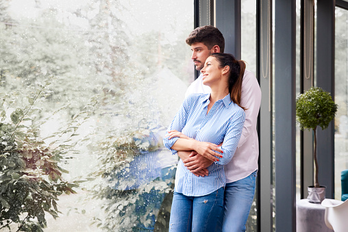 Portrait shot of happy young couple standing by the window and enjoying each otherâs company while snowing outside.