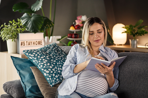 Front view, cut out of the young, blond woman in late pregnancy, sitting comfortably on the couch in her living room an making a list of baby names.