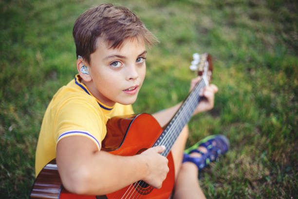 Hard of hearing preteen boy playing guitar outdoors. Child with hearing aids in ears playing music and singing song in park. Hobby art activity for children kids. Authentic childhood moment. stock photo