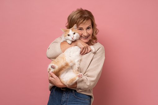 Caucasian mature woman lovingly hugging cat on pink wall.