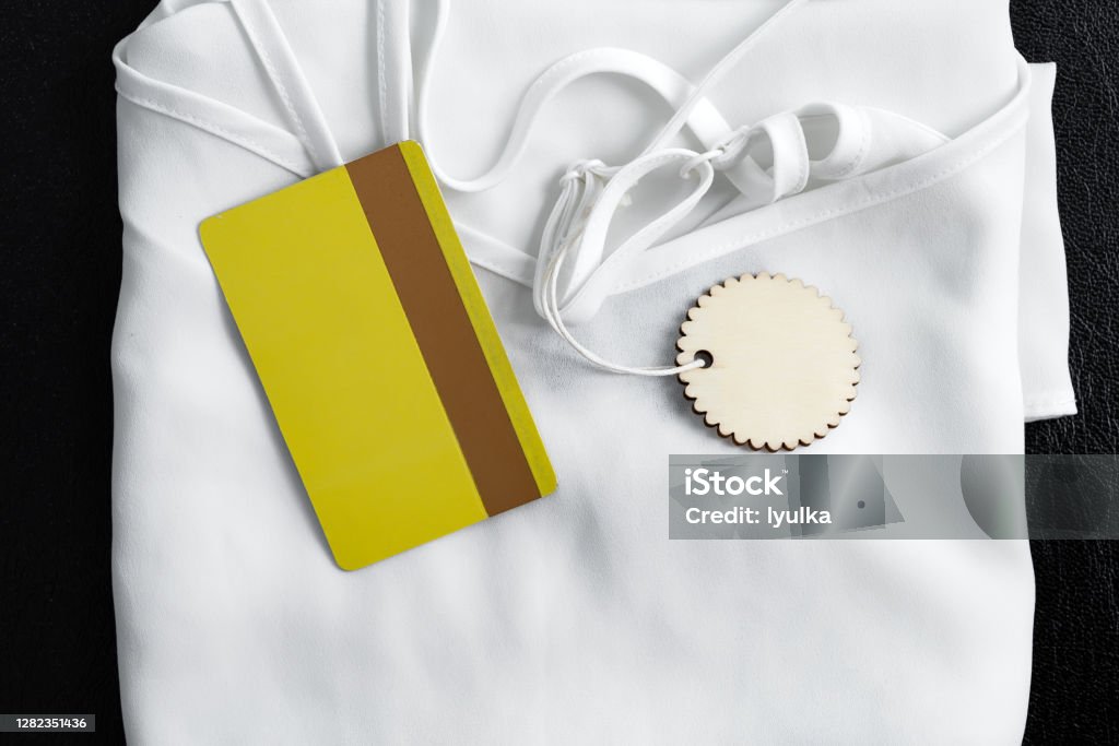 New woman clothes and gold credi card on black background. New woman white clothes with label and mock up discount or credit yellow card on a black background. Top view. Black Friday Sale concept. Advertisement Stock Photo