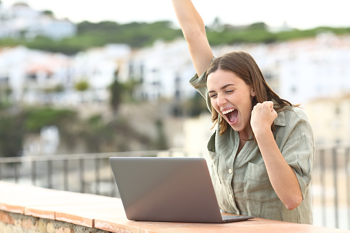 Excited woman checking laptop in a balcony in a town