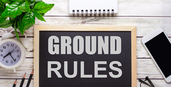 The word GROUND RULES written on a black background near pencils, a smartphone, a white notepad and a green plant in a pot