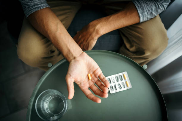 junger mann hält eine pille in der hand vor einem tisch mit einem glas wasser. medizinische behandlung / drogenkonsumkonzept. - prozac stock-fotos und bilder