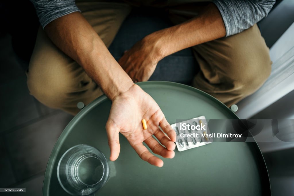 Young man holding a pill in his hand in front of a table with glass of water. Medical treatment / drug use concept. Young man holding a pill in his hand. Medical treatment / drug use concept. Illness/ health problems concept. Pill Stock Photo
