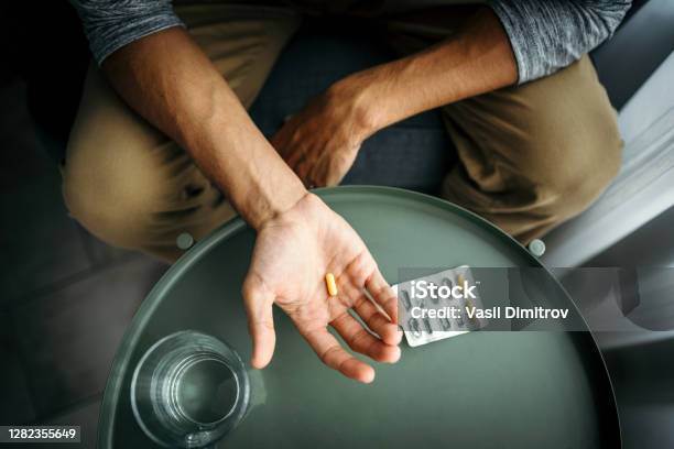 Young Man Holding A Pill In His Hand In Front Of A Table With Glass Of Water Medical Treatment Drug Use Concept Stock Photo - Download Image Now