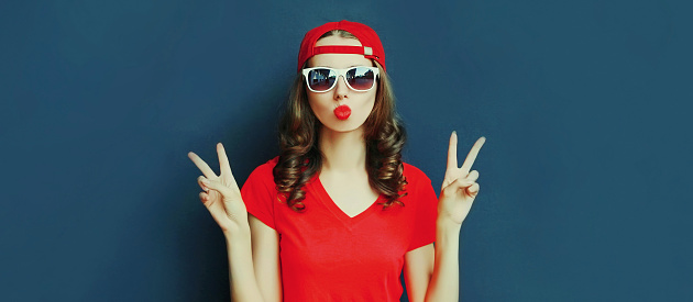 Portrait close up of young woman showing peace sign gesture wearing baseball cap over blue background