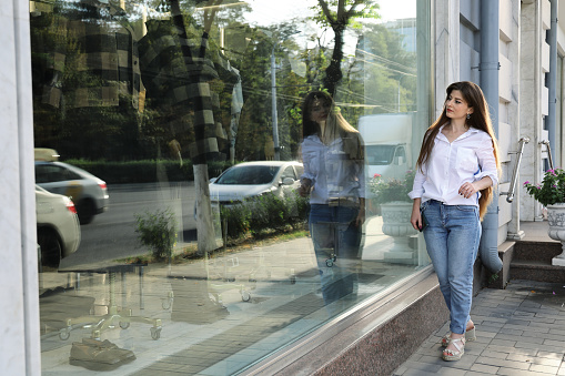 Young beautiful woman in casual style stands outside and looks into shop window with branded clothes