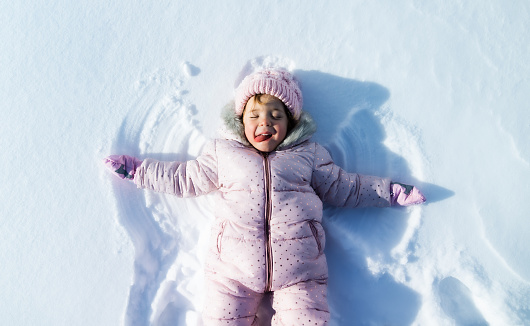 Top view portrait of cheerful small toddler girl lying in snow in winter nature, making angels.