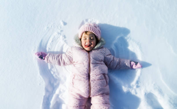 widok z góry portret wesołej małej dziewczynki leżącej w śniegu w zimowej przyrodzie, robiącej anioły. - hat toddler little girls pink zdjęcia i obrazy z banku zdjęć