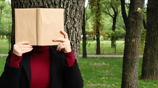 Portrait of a young woman with an open untitled book held close to her face. Brunette covering her face with a book in the park on a sunny day. Education and people concept, selective focus