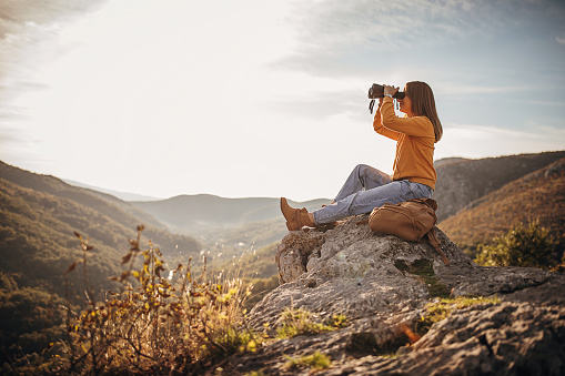 One woman, female hiker sitting on mountain peak in sunset alone, looking at the view with binoculars.