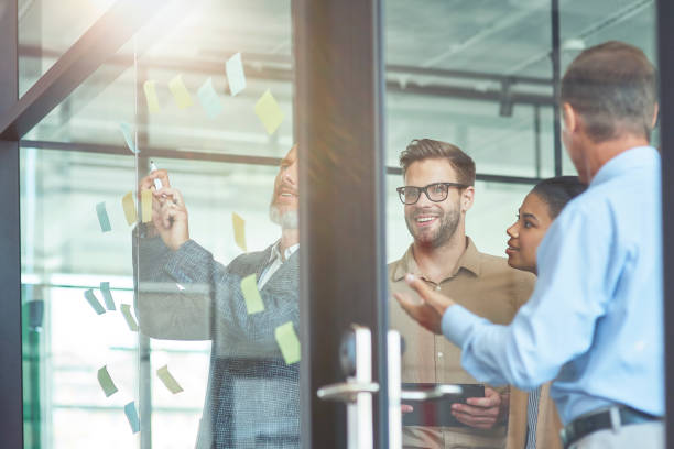 istock Group of creative coworkers putting colorful sticky notes on a glass wall, discussing something and smiling while standing in the modern office 1282372585