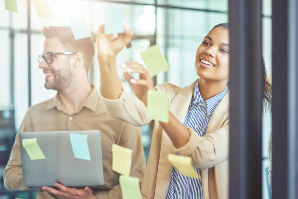istock Project planning. Two creative diverse coworkers putting colorful sticky notes on a glass wall while standing in the modern office 1282372729