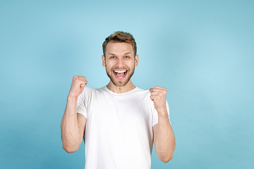 Concept of win, winner and victory. Excited and happy young adult man raised fists up, screaming loud, smiling wide, standing isolated on blue copy space background in casual white t-shirt