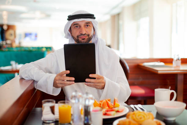 homme arabe s’asseyant à la table utilisant la tablette numérique pendant le petit déjeuner - restaurant business person setting the table clothing photos et images de collection