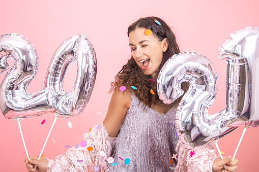 Beautiful brunette party girl with curly hair and festive clothes posing on a pink studio background with confetti and holding silver balloons from the numbers 2021 in her hand, happy new year concept
