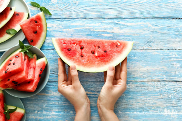 istock Fresh red watermelon slice in female hands on an blue rustic wood background with copy space. Top view. Summertime concept. Watermelon slice and mint on the background. 1282385372