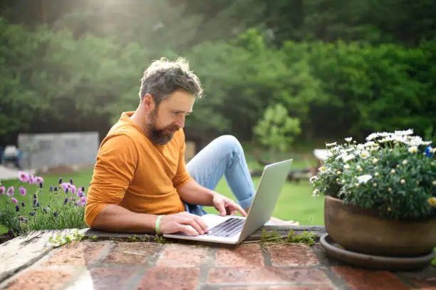 Photo of Mature man with laptop working outdoors in garden, home office concept.