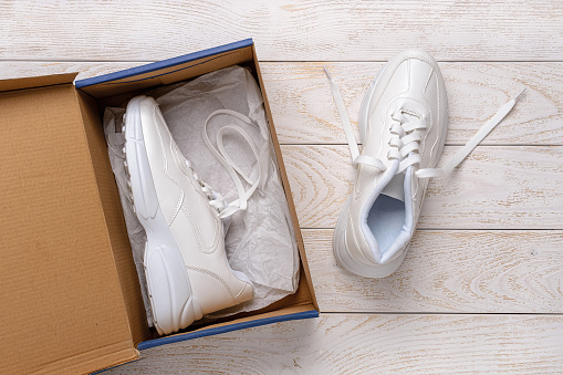 One white chunky sole sneakers in a brown cardboard box, the other on the white wood floor. Open box with new comfortable shoes for active lifestyle, fitness and sports. Top view.