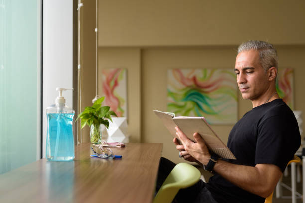 Handsome Persian man with gray hair reading book at the library Portrait of handsome Persian man with gray hair at the library inside modern building middle eastern ethnicity mature adult book reading stock pictures, royalty-free photos & images