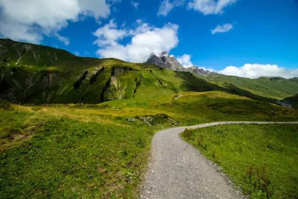 A gravel path bending rightwards in front of a grass-covered hill in the Austrian Alps. The peak in the background is the Großer Widderstein in Vorarlberg, Austria.