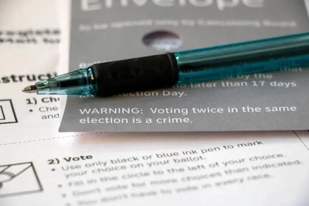 Closeup edge of a mail voting envelope on top of other voter materials, focused on text 'Warning: Voting twice in the same election is a crime' with pen laying on top.