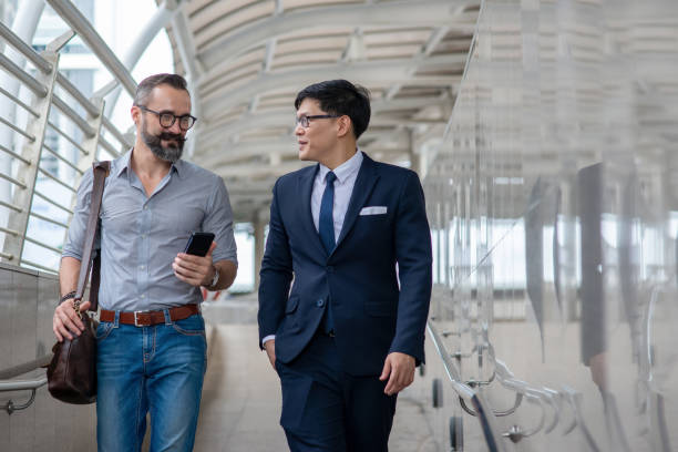 istock Two businessman walking together on office district and discussing business project with using smartphone. Caucasian beard man with Asian man office worker colleague talking about working plan 1282427757