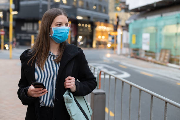 istock Young businesswoman with protective face mask using smartphone while commuting in downtown city street and city buildings with multi-coloured commercial signs at night 1282429048