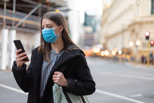 istock Young businesswoman with protective face mask using smartphone while commuting in downtown city street and city buildings with multi-coloured commercial signs at night 1282429049