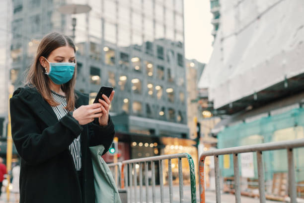 istock Young businesswoman with protective face mask using smartphone while commuting in downtown city street and city buildings with multi-coloured commercial signs at night 1282429053