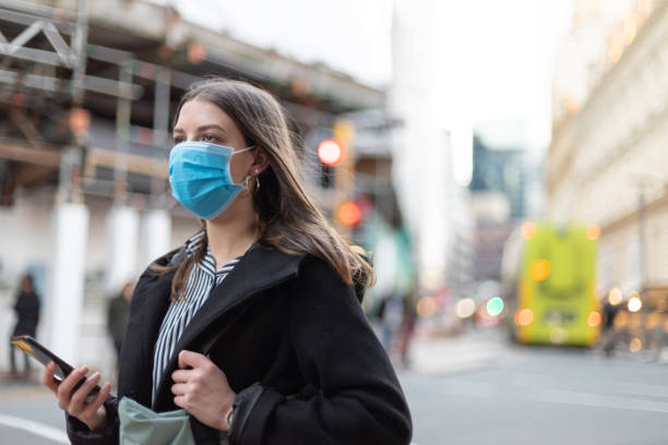 istock Young businesswoman with protective face mask using smartphone while commuting in downtown city street and city buildings with multi-coloured commercial signs at night 1282429054