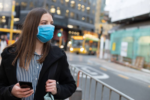 istock Young businesswoman with protective face mask using smartphone while commuting in downtown city street and city buildings with multi-coloured commercial signs at night 1282429968