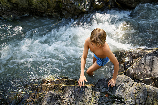 Little boy enjoying the cold mountain river. He is climbing up the stones near the strong river current.\nNikon D850