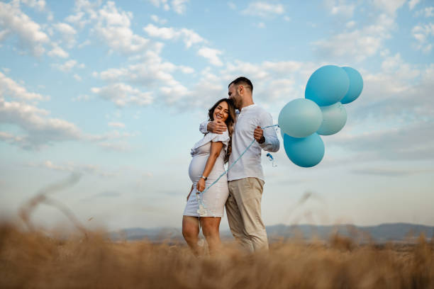 Pregnant woman and man in love holding blue balloons in nature A young pregnant woman and a young man dressed in white holding blue balloons in nature on a sunny summer day couple hope sky blue stock pictures, royalty-free photos & images