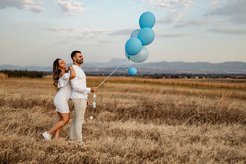 A young pregnant woman and a young man dressed in white holding blue balloons in nature on a sunny summer day
