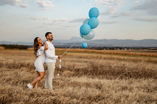 Pregnant woman and man with blue balloons in nature A young pregnant woman and a young man dressed in white holding blue balloons in nature on a sunny summer day couple hope sky blue stock pictures, royalty-free photos & images