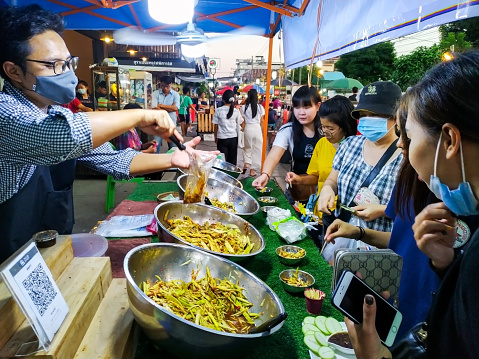 Kad Kong Ta, Lampang, Thailand.- Oct25, 2020 : man market vendors with face mask, putting mango salad in plastic bag for selling , with people waiting for queue  at night market on october25, 2020.