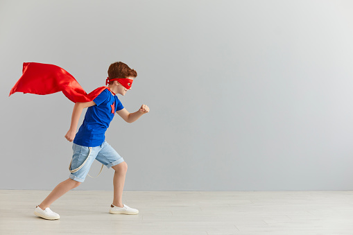 Little boy in a superhero costume with a red cloak runs towards the free space for text on a background of a gray wall. Child of a superhero. Place for text or advertising.