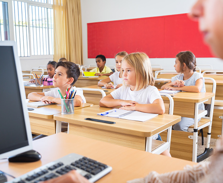 Diligent tweens studying in classroom, listening to female teacher and writing in notebooks