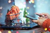 The girl presses her hand on the smartphone screen and holds a credit card in her hands. Trolley with gifts on the background of a laptop. Online shopping from home. Christmas and New Year sales
