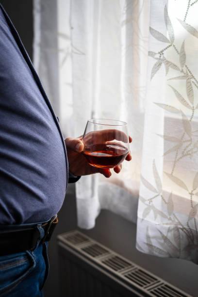 homme en surpoids avec un verre de cognac dans la main à une fenêtre - abdomen addiction adult alcohol photos et images de collection