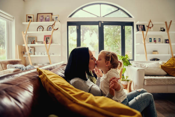 Playful young woman kissing toddler girl while sitting on sofa at home Mother hugging and kissing daughter sitting on couch mouths kissing stock pictures, royalty-free photos & images