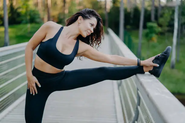 Sporty slim brunette woman dressed in cropped top and leggings stretches legs on bridge does warm up before morning run pose outdoor wants to have slim body and good health. Workout, lifestyle