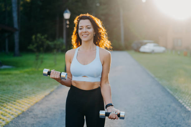 Photo of slim brunette young woman raises dumbbells has morning workout poses against sunrise dressed in cropped top and leggings works on arms muscles smiles positively leads sporty lifestyle ripl fitness
