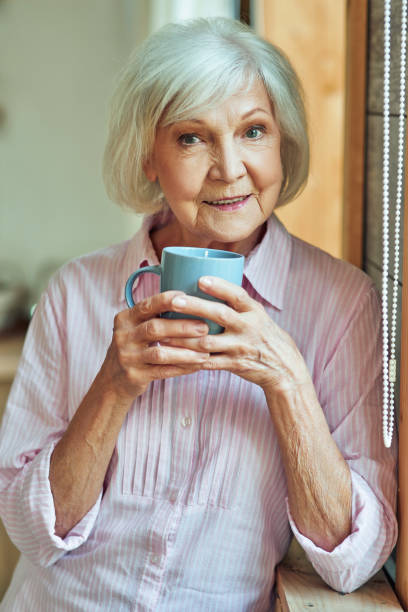 istock Happy stylish gray haired lady standing near window with cup 1282480183