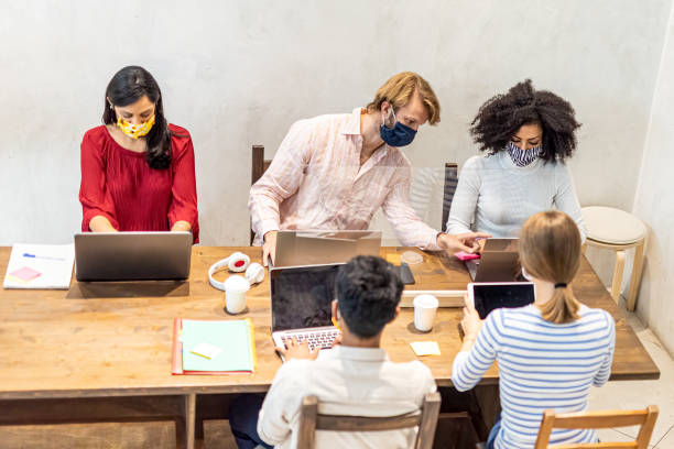 istock Multiethnic group of co-workers at work in a temporary office, protective face masks worn, plexiglass barrier to protect health, the new normal concept applied to smart business 1282481767
