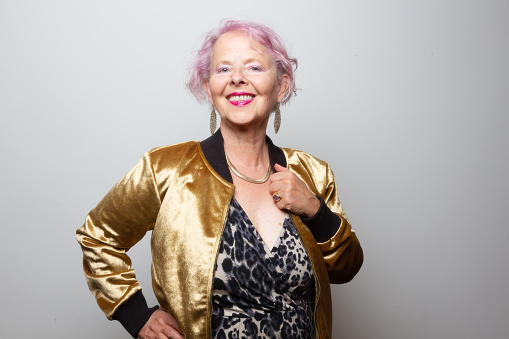 Senior woman with dyed pink hair, 65 years old, wearing dress with leopard print pattern and a golden jacket, a well as huge golden jewellery,  looking to camera with a toothy smile