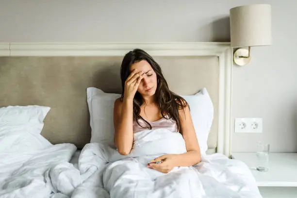 Young caucasian woman lying in bed, feeling unwell, with her hand on her head. Sickness / illness concept. Coronavirus / fever / headache concept. Home isolation.