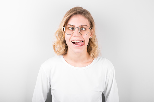 Blonde attractive female makes grimace, foolishes in studio, crosses eyes and rounds lips, has fun after being tired at classes, isolated over white background.